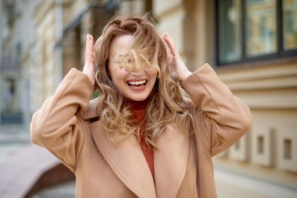 smiling woman walk in center and protect hair from wind gust at street. girl hands straighten hair